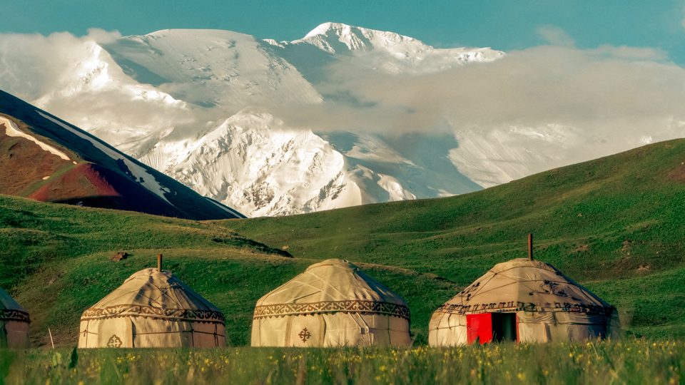 Yurts on a green slope in front of snowy Trans-Alay peaks.