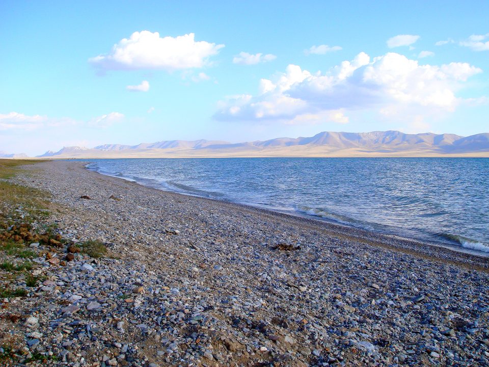 Stony shore and open water of Son-Kul Lake with mountains in the background.